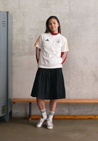 Νεαρή γυναίκα στέκεται δίπλα σε ντουλάπια και ένα παγκάκι, φορώντας μια λευκή αθλητική φανέλα πάνω από ένα κόκκινο μακρυμάνικο μπλουζάκι, μαύρη φούστα, ριγέ καλτσόν και αθλητικά παπούτσια.