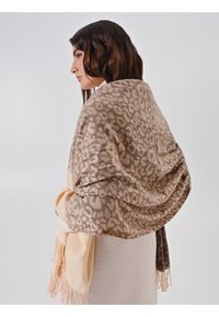 Femme avec des cheveux brun moyen portant un châle léopard beidge et marron sur un manteau clair, de profil à gauche sur un fond uni.