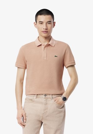 Camiseta polo melocotón hecha de algodón, con cuello, mangas cortas y un pequeño logo verde. Combinada con jeans de color claro y un reloj.