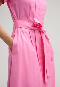Rosa Kleid mit taillierter Oberseite, plissiertem Rock und einem gebundenen Gürtel in der Taille. Hergestellt aus glattem Stoff mit Seitentaschen.