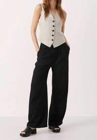 Beige-rivinen liivi V-pääntiellä ja viidellä napilla, yhdistettynä korkeavyötäröisiin, laskospöksyihin, joissa on mustat leveälahkeiset housut, sekä mustiin liukusandaleihin, joissa on niittejä.