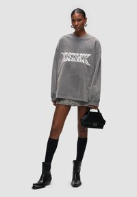 Felpa grigia oversize con testo grafico bianco, gonna mini a fantasia, borsa a tracolla nera, calze alte nere fino al ginocchio e stivaletti neri con tacco.