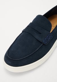 Chaussure slip-on en daim marine avec un bout arrondi, dotée d'une sangle décorative et d'un détail de couture bleue. Semelle en caoutchouc blanche.