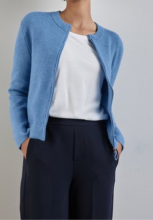 Personne portant un cardigan bleu clair zippé sur un t-shirt blanc avec un pantalon noir, les mains dans les poches, debout contre un mur uni.