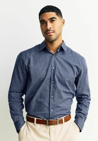 Koszula zapinana na guziki w granatowo-biały wzór w kratę, z długimi rękawami i kontrastowymi guzikami, zestawiona z brązowym paskiem i jasnymi spodniami.