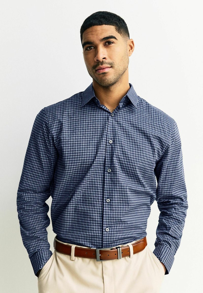 Koszula zapinana na guziki w granatowo-biały wzór w kratę, z długimi rękawami i kontrastowymi guzikami, zestawiona z brązowym paskiem i jasnymi spodniami.