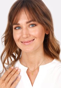 Collier en or avec un petit pendentif ovale, porté par une personne aux cheveux bruns ondulés, sur un fond uni.