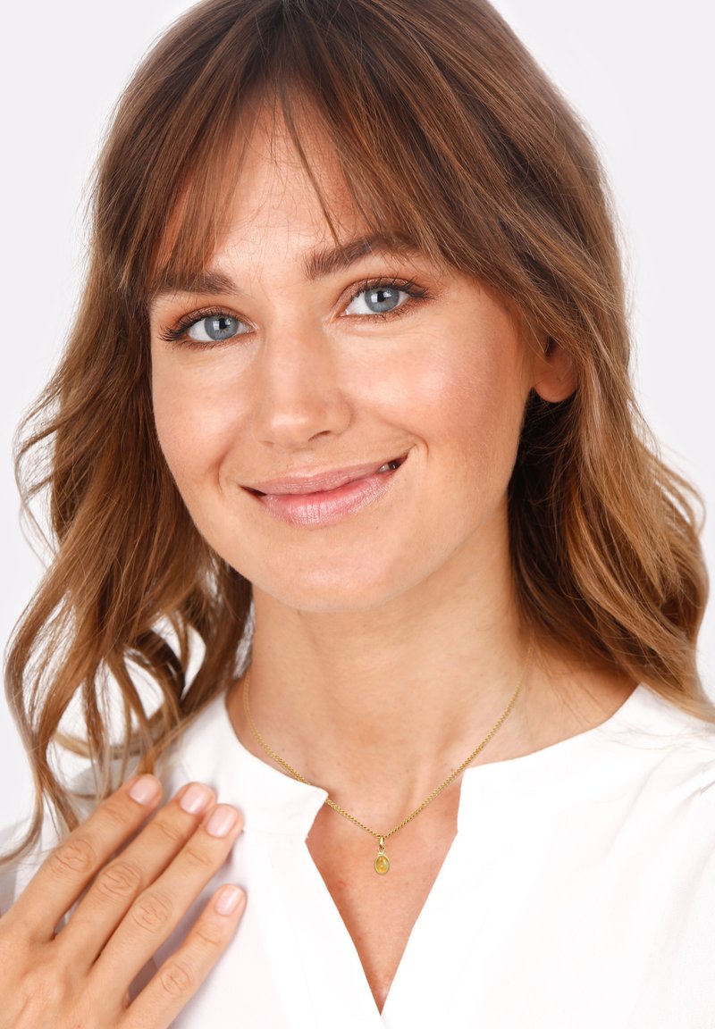 Collier en or avec un petit pendentif ovale, porté par une personne aux cheveux bruns ondulés, sur un fond uni.