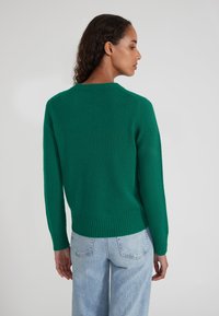 Groene gebreide trui met een ronde hals, geribde manchetten en zoom; gladde textuur; gedragen met lichtblauwe high-waisted jeans.