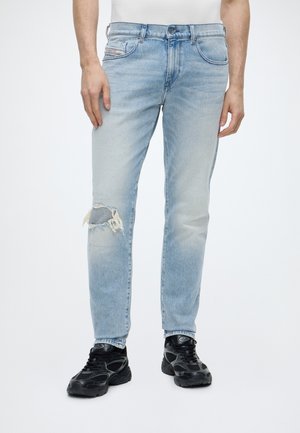 Homme portant un jean bleu clair déchiré au genou gauche et des baskets sportives noires, debout devant un fond blanc.