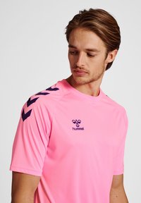 Camisa deportiva rosa hecha de un tejido suave y que absorbe la humedad. Presenta un cuello redondo, un logotipo morado y patrones de chevron morado en contraste en las mangas.