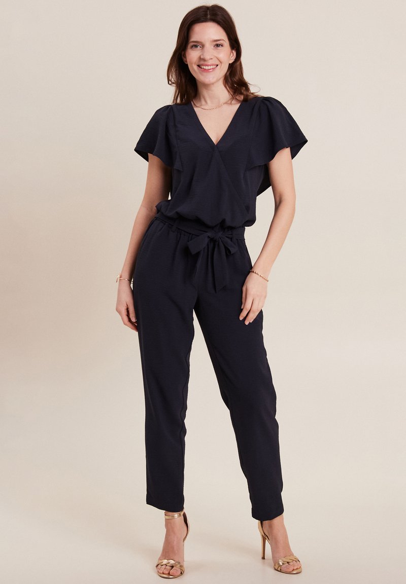 Tuta navy con maniche corte a sbuffo, scollatura a V, vita con nodo e gambe affusolate. Presenta una texture leggera e una vestibilità rilassata.
