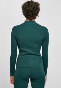 Türkisfarbenes, geripptes Langarmshirt mit hohem Kragen, das eine figurbetonte Silhouette und strukturierte Stoffstruktur aufweist. Rückansicht zeigt glatte Nähte und leichte Dehnbarkeit.