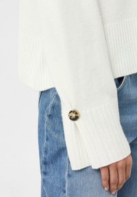 Weißer, gerippter Pullover mit weitem, offenem Bündchen, das einen runden, schildpattfarbenen Knopf hat, kombiniert mit hellblauen Jeans.