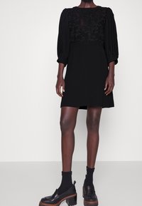 Černé šaty s nabíranými rukávy, květinovou krajkovou korzetou a rovnými liniemi. Nosené s černými ponožkami a platformovými mokasíny. Hladký vzhled materiálu.