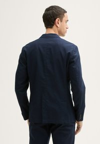 Sacou bleumarin dintr-un material texturat, având un singur slit, rever în formă de vârf și manșete cu nasturi. Croială ajustată cu linii curate.
