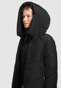 Chaqueta acolchada negra con una capucha grande y estructurada. Cuenta con un cierre de cremallera, botones a presión y un bolsillo exterior con una textura sutil.