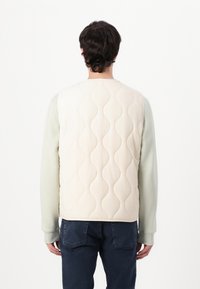 Chaleco acolchado color crema con patrón de olas, hecho de un tejido suave. Combinado con una camisa de manga larga de color verde claro y jeans azul oscuro.