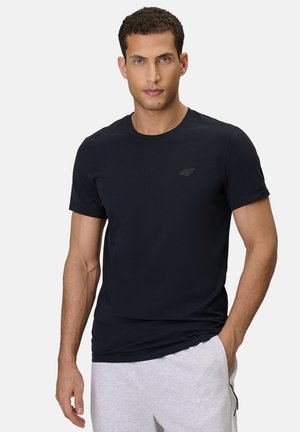 Mężczyzna o krótkich, kręconych włosach, ubrany w czarny t-shirt z logo 4F i jasnoszare spodnie dresowe, stoi z jedną ręką w kieszeni.