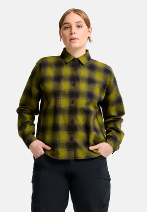 Persona che indossa una camicia a quadri verde e nera abbottonata, con le mani nelle tasche e pantaloni neri, in piedi davanti a uno sfondo bianco semplice.