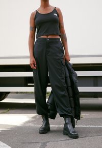 Top corto negro con logo, pantalones cargo anchos negros y botas de tobillo negras con suelas gruesas, sosteniendo una chaqueta acolchada negra.