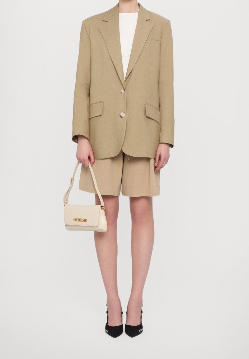 Beige oversize blazer, ljus topp, beige shorts, svarta klackar, håller en krämfärgad handväska med "LOVE MOSCHINO" i guldtext.