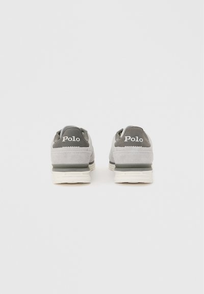 Ζευγάρι γκρι αθλητικά παπούτσια Polo, εμφανιζόμενα από πίσω σε λευκό φόντο, με λευκές σόλες και γκρι πίσω πάνελ με την επιγραφή "Polo".