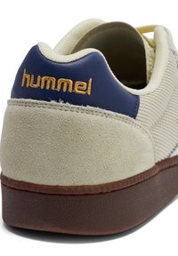 Svetlo bež superga z mornarsko modrim petnim delom, zgornji del iz semiša in blaga, gumijast podplat ter z vezenim zlatim logotipom "hummel" na zadnji strani.