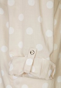 Blusa beige con pois bianchi, caratterizzata da un polsino piegato con un bottone argentato. Tessuto leggero con una texture morbida e liscia.