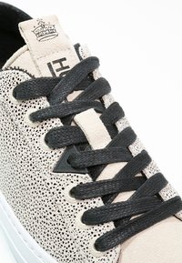 Gros plan d'une basket beige avec des éclats noirs, des lacets noirs et un logo de couronne cousu sur la languette.