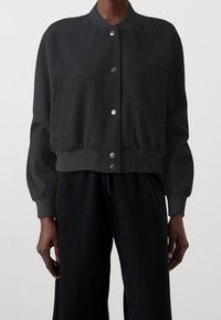 Svart kort bomberjacka i mjukt material, med rund halsringning, två stora framfickor och metallknappar.