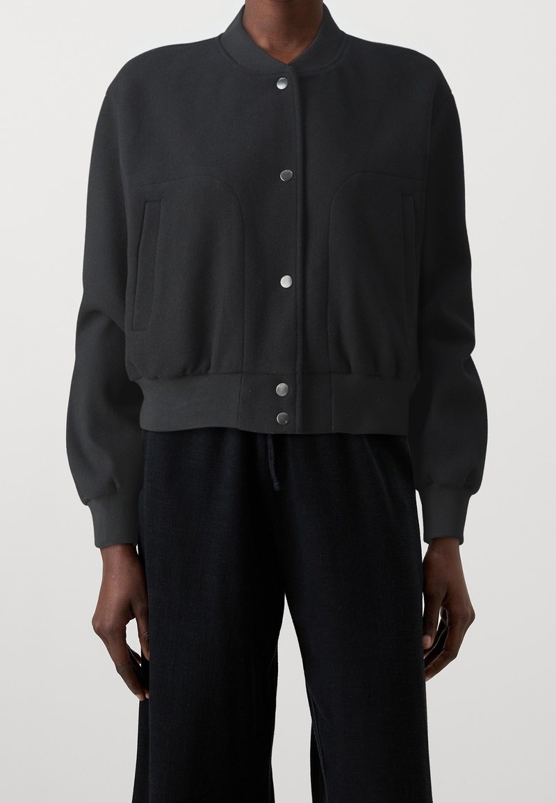 Svart kort bomberjacka i mjukt material, med rund halsringning, två stora framfickor och metallknappar.