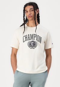 Gaiši krēmveida T-krekls ar īsām piedurknēm, kam ir izteiksmīgs melns "CHAMPION" logo un apaļa emblema ar lauriem zem tā.