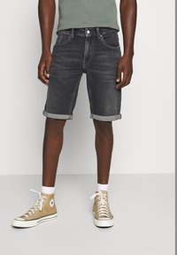 Tummansiniset farkkuhousut lyhyinä, käännetyt lahkeet, yhdistettynä vihreään paitaan ja beigeihin korkeisiin tennareihin, joissa on valkoiset nauhat ja kumipohjat.