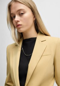 Yellow tailored blazer with notch lapels, single chest pocket; worn over a black crew neck top, featuring a silver chain necklace.