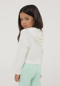 Felpa bianca corta con cappuccio, polsini e vita elastici, abbinata a leggings verde menta, creando un look casual.