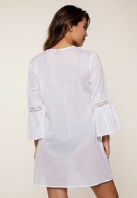 Vestido blanco ligero con mangas largas acampanadas y detalles de encaje. La tela es ligeramente translúcida, con un corte holgado y un escote redondo.