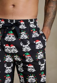 Fekete pizsama rövidnadrág, amely ismétlődő mintázatban ábrázolja a rohamosztagos sisakokat, melyeken mikulássapkák, agancsok és színes fények díszítik, húzózsinóros derékrésszel.