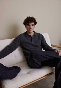 Grijze, textuur geverfde langemouwen polo gedragen door een persoon die op een witte, stoffeerder sofa zit, met donkere streepjesbroek, tegen een effen achtergrond.