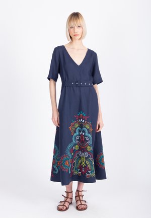 Kobieta w granatowej sukience midi z dekoltem w szpic, krótkimi rękawami, paskiem i kolorowym kwiatowym haftem na dole spódnicy, nosząca brązowe sandały na paski.