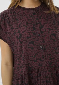 Robe à manches courtes en bordeaux foncé avec un motif floral. Dotée d'un col rond et d'une fermeture à boutons sur le devant. Détail de pliures à l'ourlet.