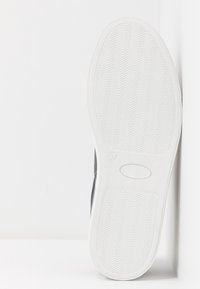 Semelle d'une chaussure, caoutchouc blanc avec une texture ondulée, affichant l'étiquette de taille "37". Partie supérieure visible sur un fond blanc, sans détails apparents.