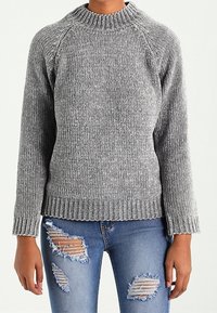 Grauer gerippter Strickpullover mit hohem Kragen und Raglanärmeln, kombiniert mit zerrissenen blauen Jeans, die mehrere Risse und ausgefranste Säume aufweisen.