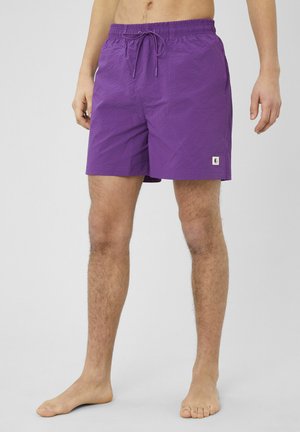 Homme portant un short de bain violet avec taille à cordon, debout pieds nus devant un fond gris clair uni.
