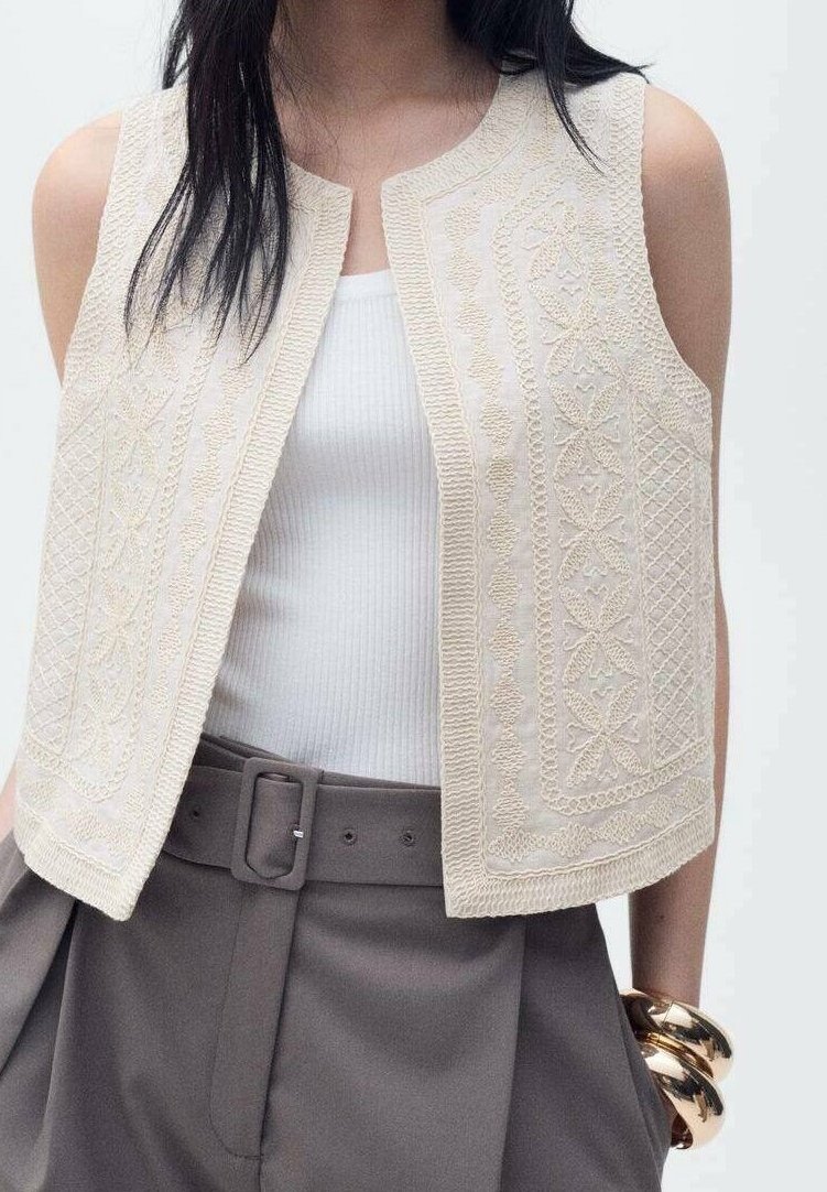 Femme portant un gilet brodé crème sans manches sur un débardeur blanc côtelé, un pantalon gris ceinturé et des bracelets épais dorés.
