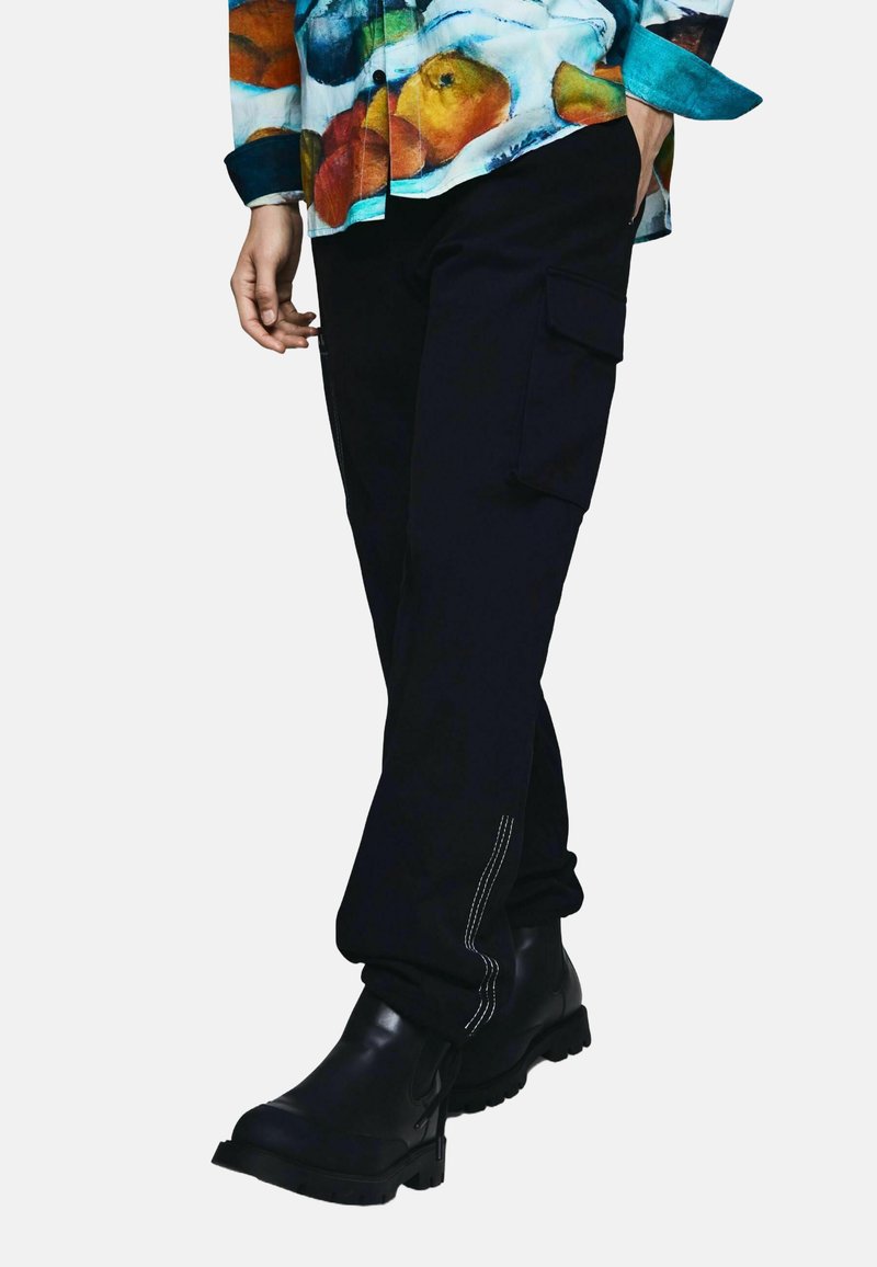 Pantalon cargo noir avec poches latérales et coutures blanches, assorti à des bottines noires. Le haut présente une chemise colorée à motif de fruits.