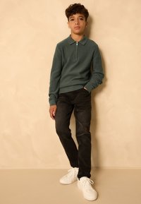 Zelený žebrovaný svetr na zip s límcem, v kombinaci s černými džíny a bílými teniskami. Oblečení je neformální a přiléhavé.