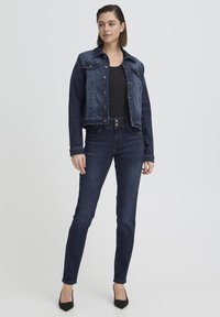 Mujer de pie con chaqueta y jeans de mezclilla azul oscuro, combinados con top negro y tacones negros, sobre un fondo claro y liso.