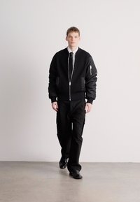 Casaco bomber preto com textura canelada, fecho de correr e bolsos laterais. Combinado com calças cargo pretas e sapatos. Modelo está em pé contra um fundo simples.