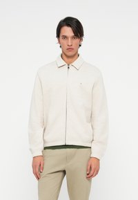 Jasno beżowa kurtka zapinana na zamek z kołnierzem, elastycznymi mankietami i pasem w talii. Zawiera małe logo na lewej piersi.